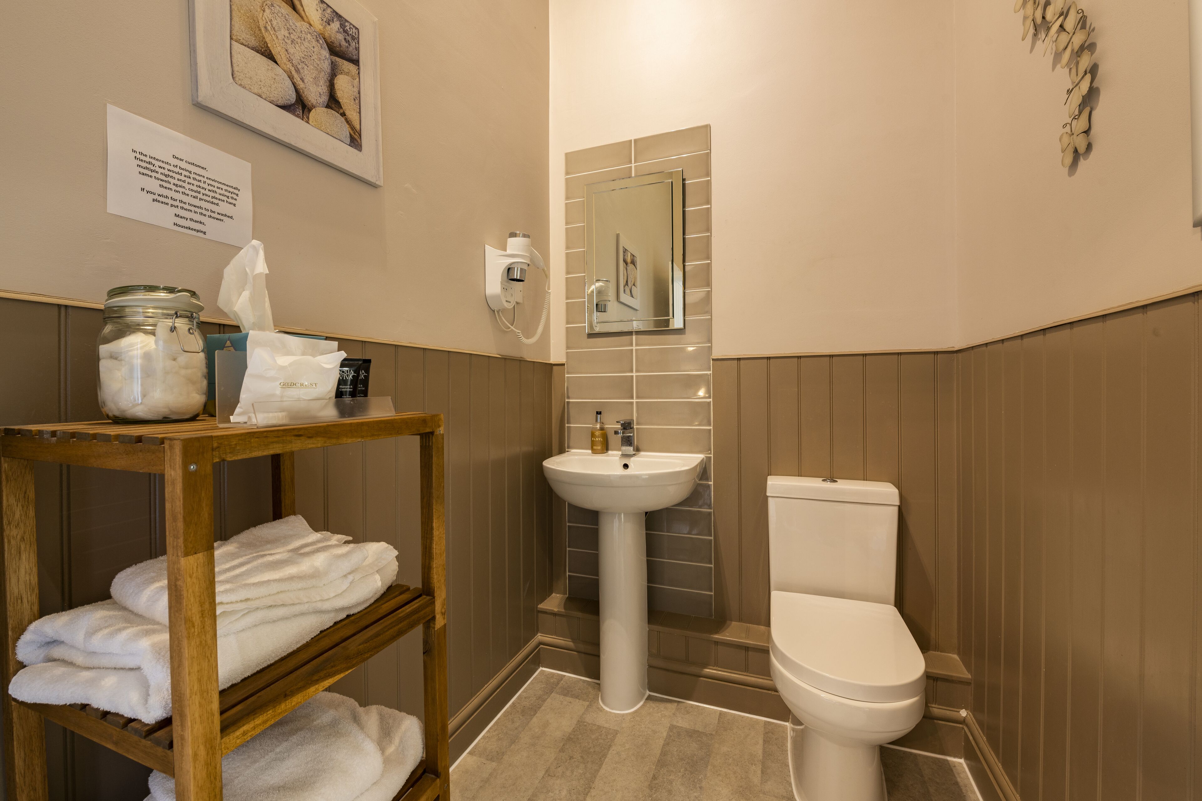 A bathroom with a white sink, a toilet, a wooden shelf with towels, a jar of cotton balls, and a framed picture on the wall.