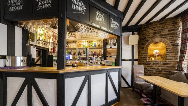 A bar area with a wooden counter, shelves of bottles, and glasses. There is a fireplace and a table with chairs. - The Swan Hotel (Wotton-under-Edge)