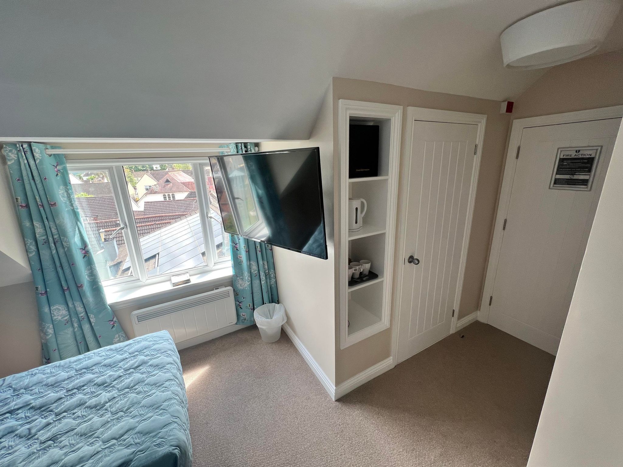 Ein Schlafzimmer mit Bett, einem Fenster mit Vorhängen, einem an der Wand montierten Fernseher, einem Kleiderschrank und einer Tür mit Brandschutzschild.
