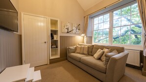 A cozy living room with a beige sofa, a small white table, and a TV mounted on the wall. There is a window with curtains and a radiator below it. - The Swan Hotel (Wotton-under-Edge)