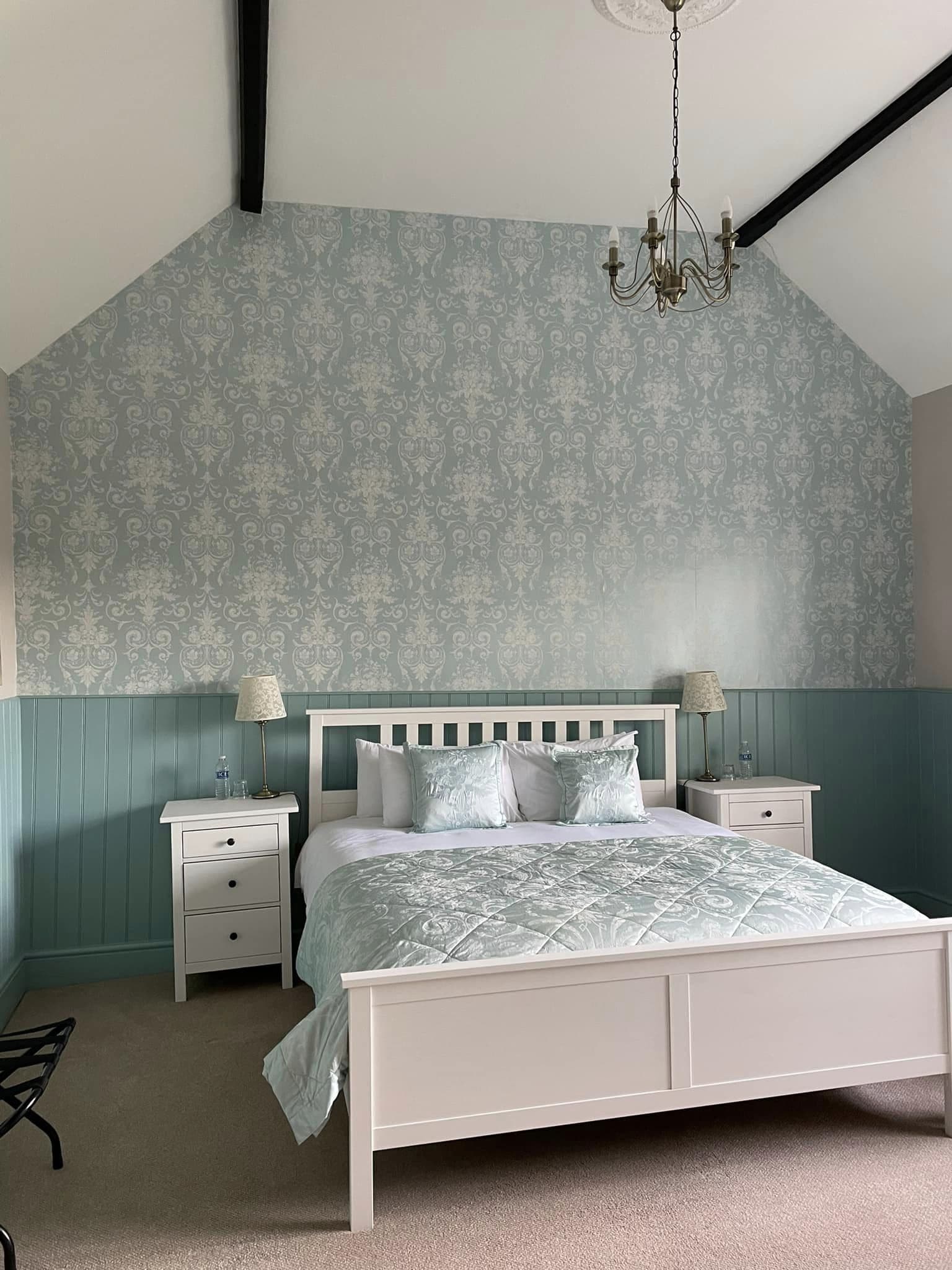 A bedroom with a white bed, two bedside tables, a chandelier, and patterned wallpaper.