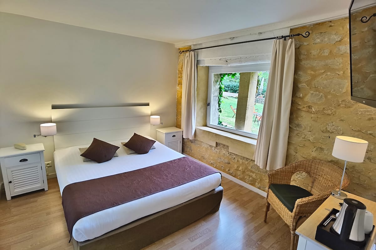 Un dormitorio con una cama grande, mesitas de noche, una silla de mimbre, una ventana con cortinas y una pared de piedra.