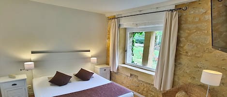 A bedroom with a large bed, bedside tables, a wicker chair, a window with curtains, and a stone wall.