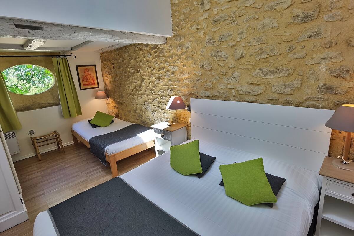 Un dormitorio con pared de piedra, dos camas, almohadas verdes y una ventana con vistas.