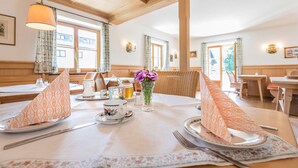 A dining area with tables set for two, featuring patterned tablecloths, floral centerpieces, and wooden beams on the ceiling. - Hotel Garni Gästehaus Brand (Bad Wiessee)