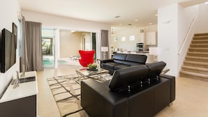 Ein modernes Wohnzimmer mit einer schwarzen Ledersofa, einem Glastisch und einem roten Sessel. Im Hintergrund ist eine Küche mit weißen Schränken und eine Treppe zu einer oberen Ebene zu sehen.