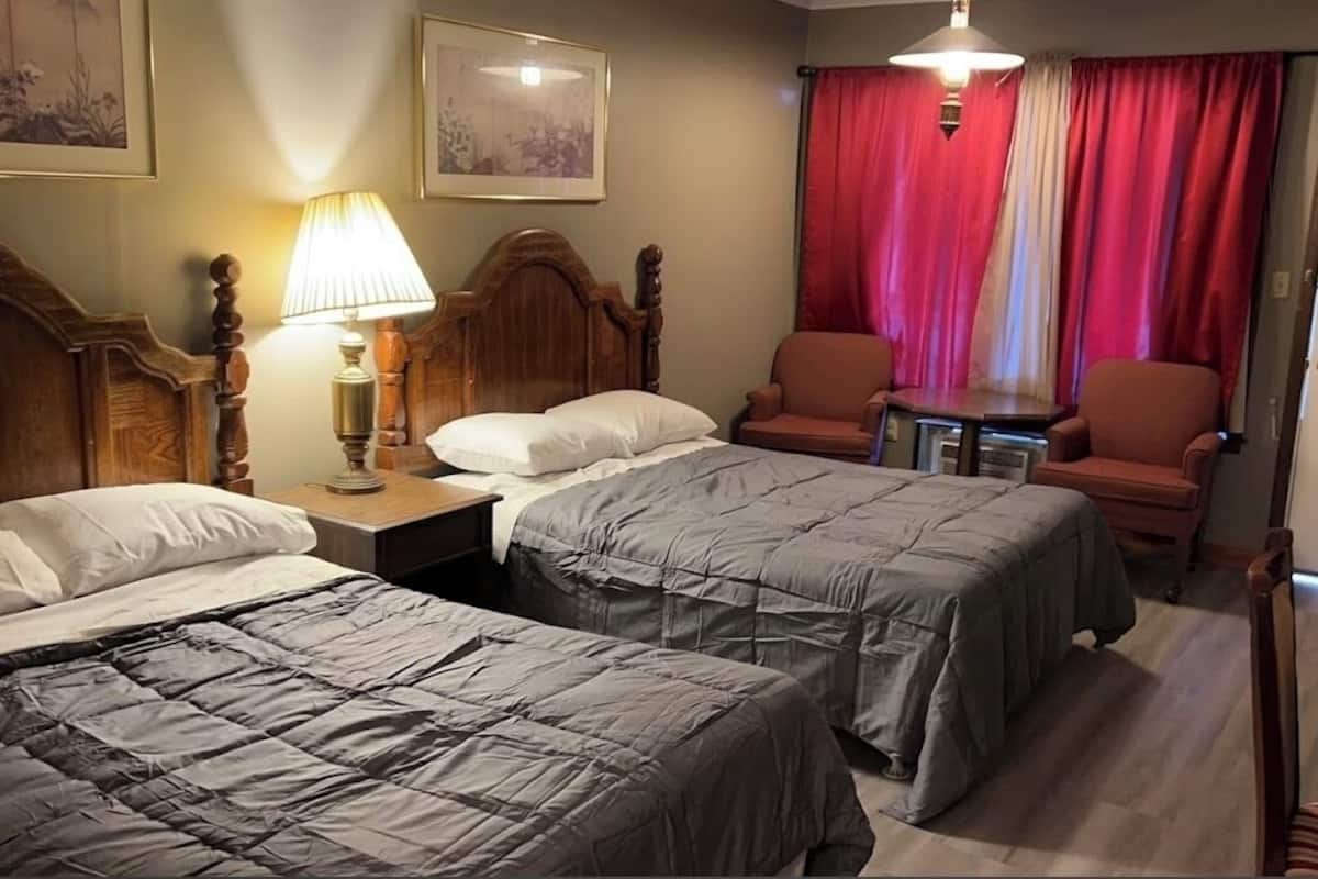 A hotel room with two beds, a nightstand with a lamp, a chair, and a small table.