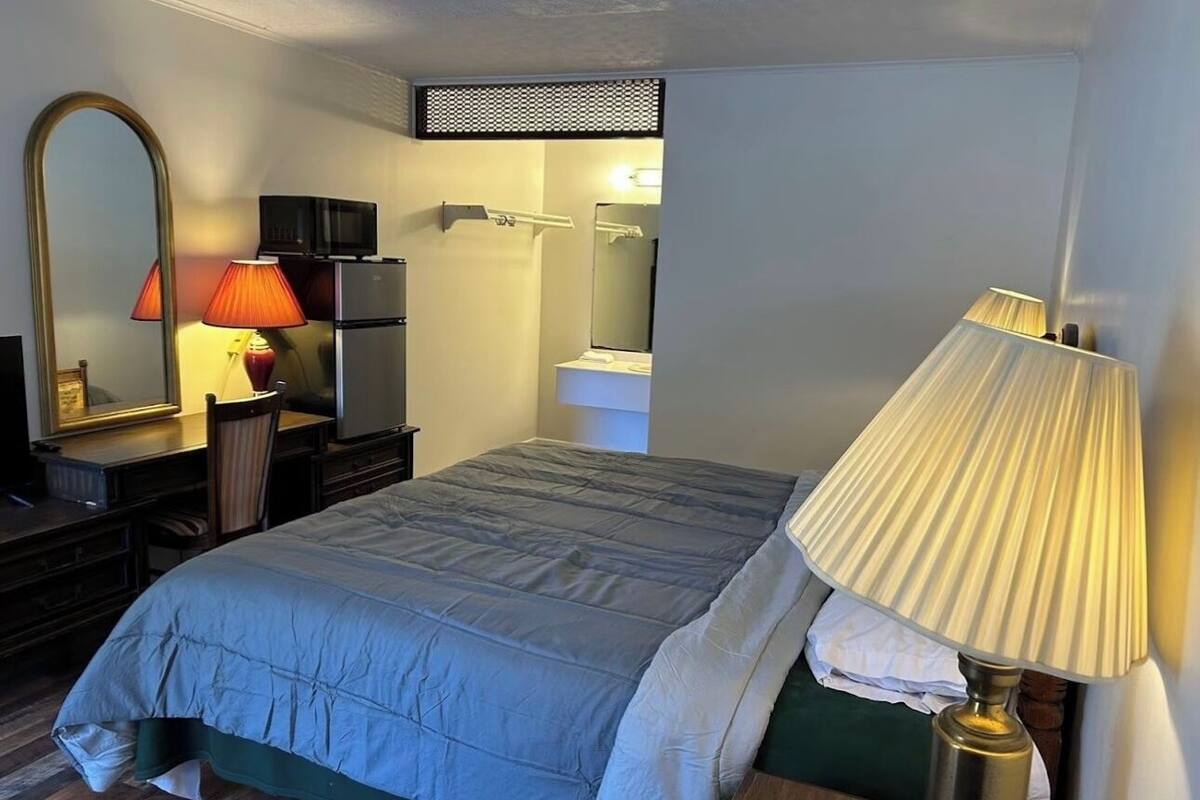 A hotel room with a bed, a desk with a lamp, a mirror, a TV, and a small bathroom area.