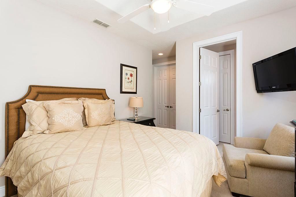 A bedroom with a large bed, a bedside table with a lamp, a TV mounted on the wall, and a door leading to another room.