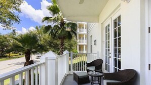 Un balcon agrémenté de meubles en osier, d’un palmier et offrant une vue sur une rue et un immeuble.