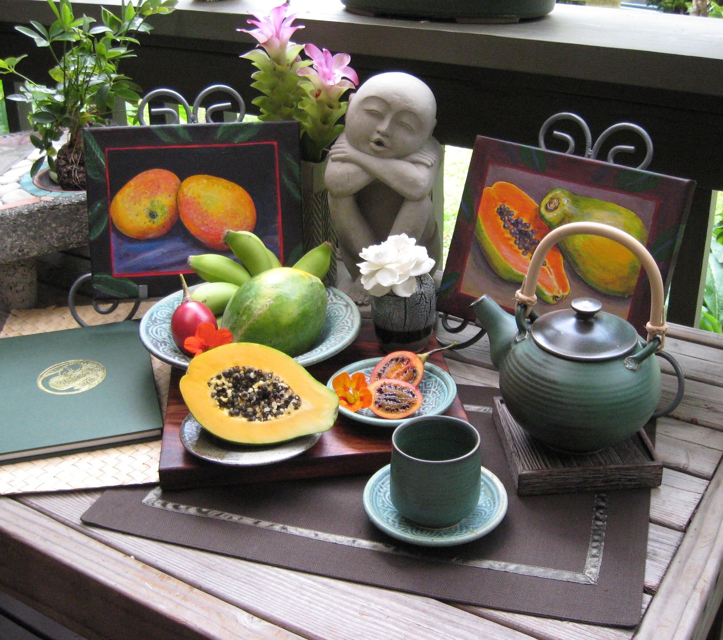 A table with a teapot, a plate with fruits, a book, and a statue.