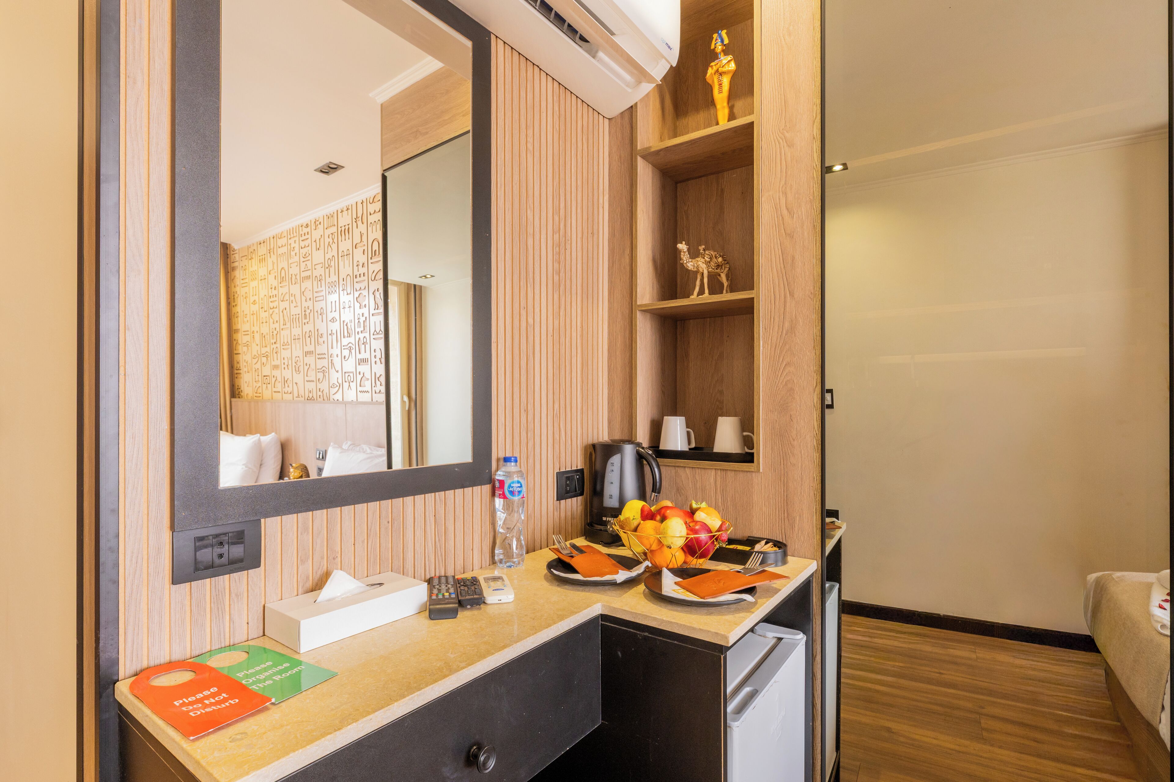 Une chambre d’hôtel avec une coiffeuse en bois, un miroir, une machine à café, un panier de fruits et un lit.