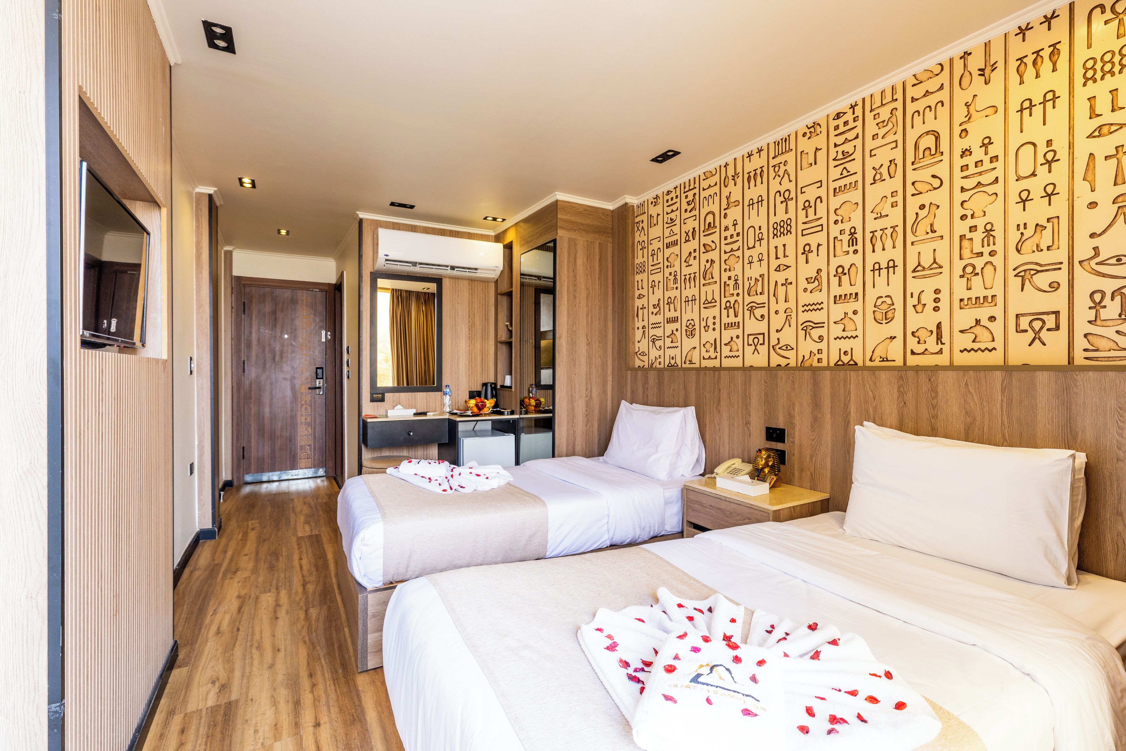 A hotel room with two beds, a wooden floor, a wall with hieroglyphs, a mirror, and a desk with a phone.
