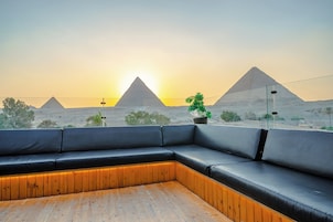 A modern lounge with a wooden floor and black leather seating, overlooking a desert landscape with pyramids during sunset.