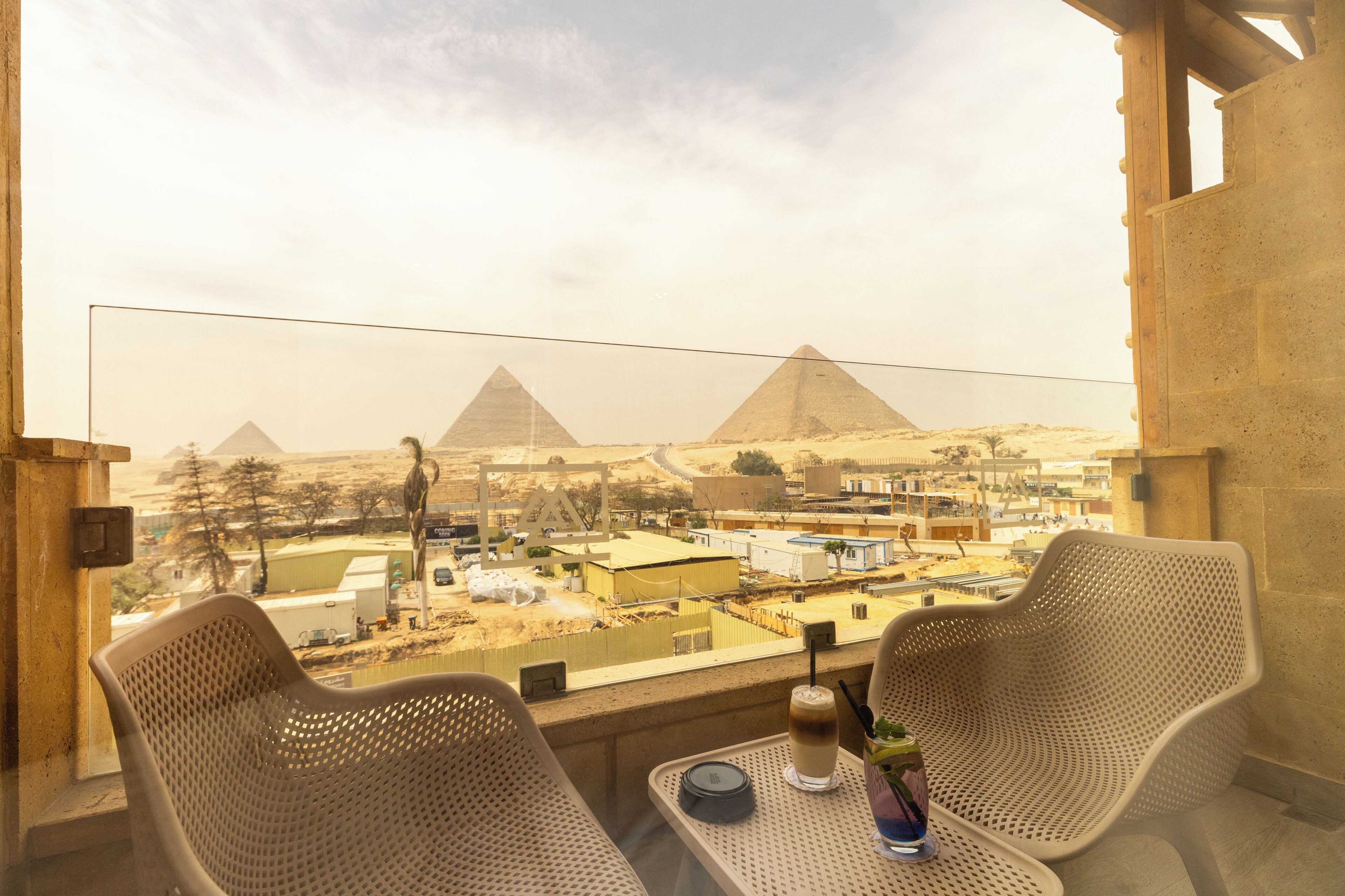 En balkon med to fletstole, et bord med drik og en flaske, og udsigt til pyramiderne.