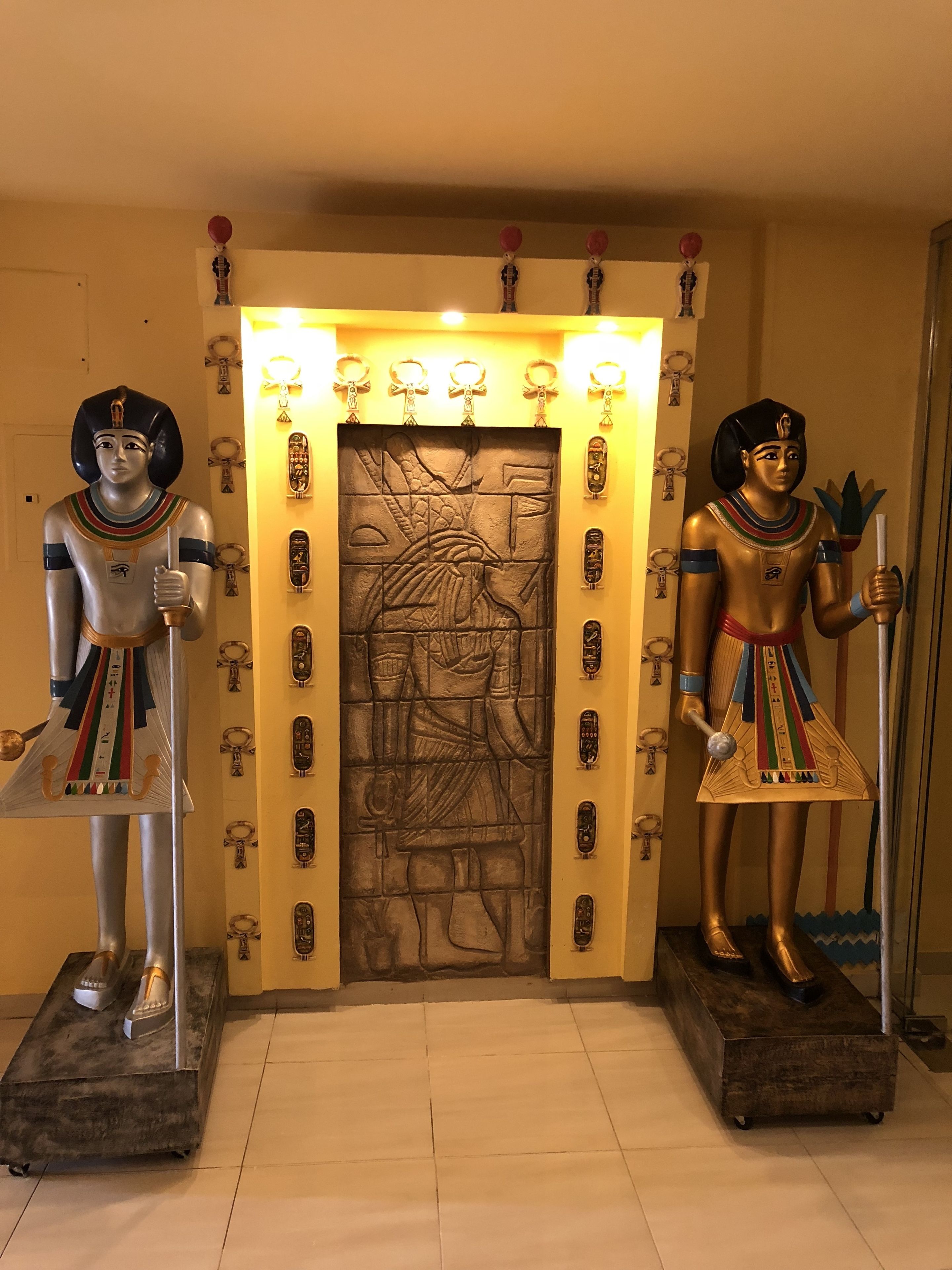 Dos estatuas de figuras egipcias frente a una pared con jeroglíficos y una puerta.