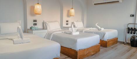 A hotel room with three beds, each with white linens and wooden headboards. There are hanging wicker lamps, an air conditioning unit, and a small table with a lamp.
