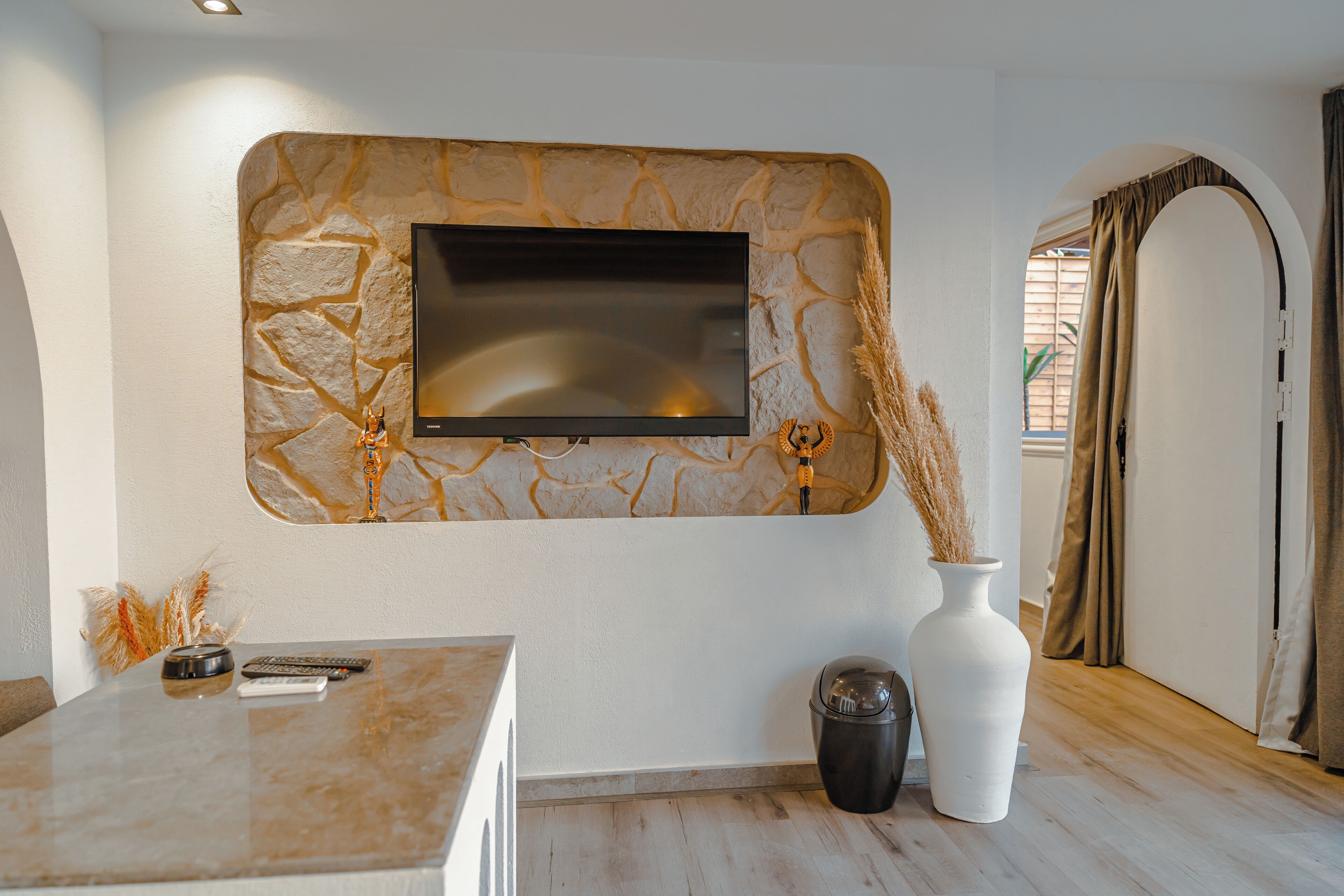 Una sala de estar moderna con una pared de piedra, un televisor de pantalla plana, un jarrón blanco y una mesita con controles remotos.