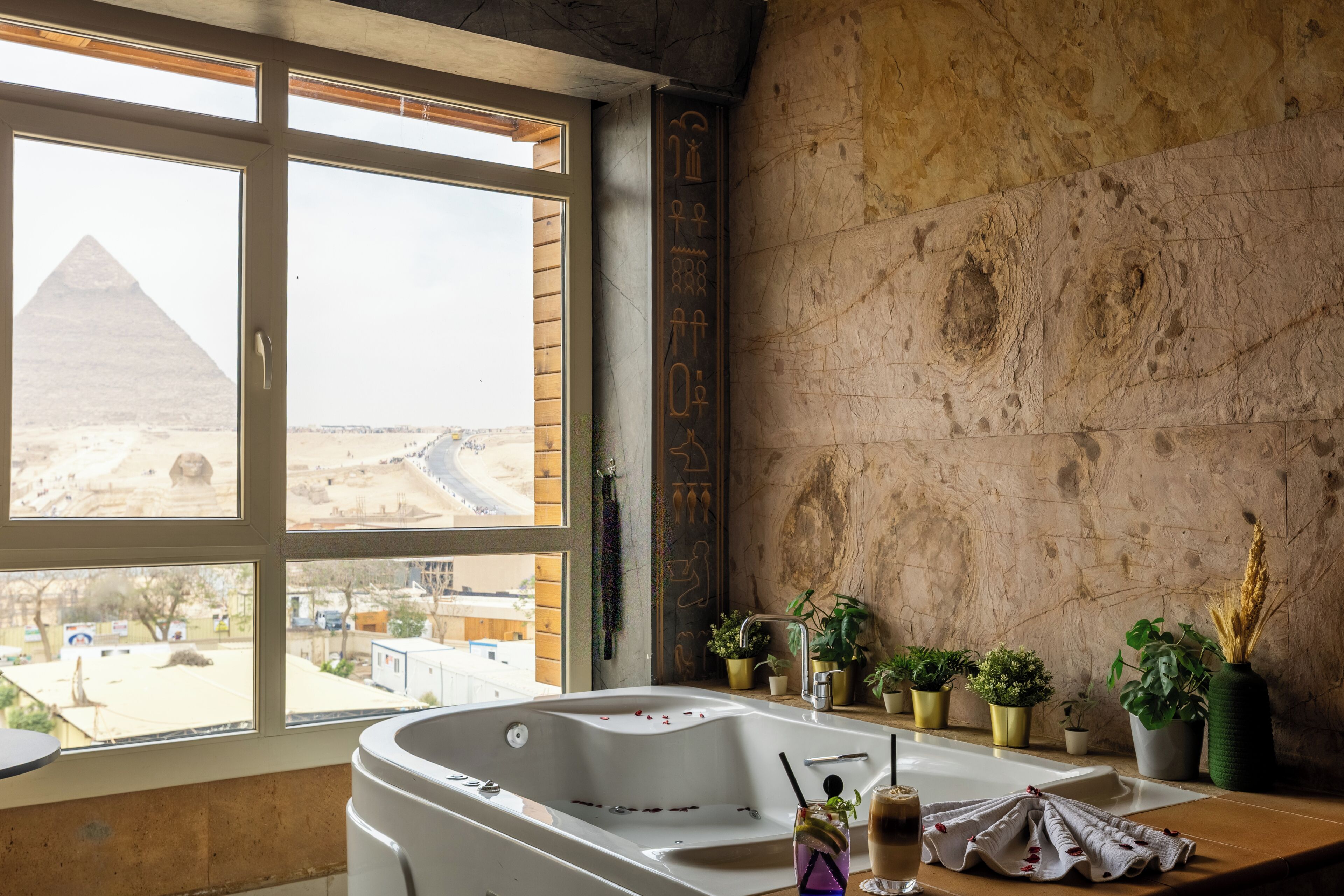 A bathroom with a bathtub, a window with a view of the pyramids, and several potted plants.