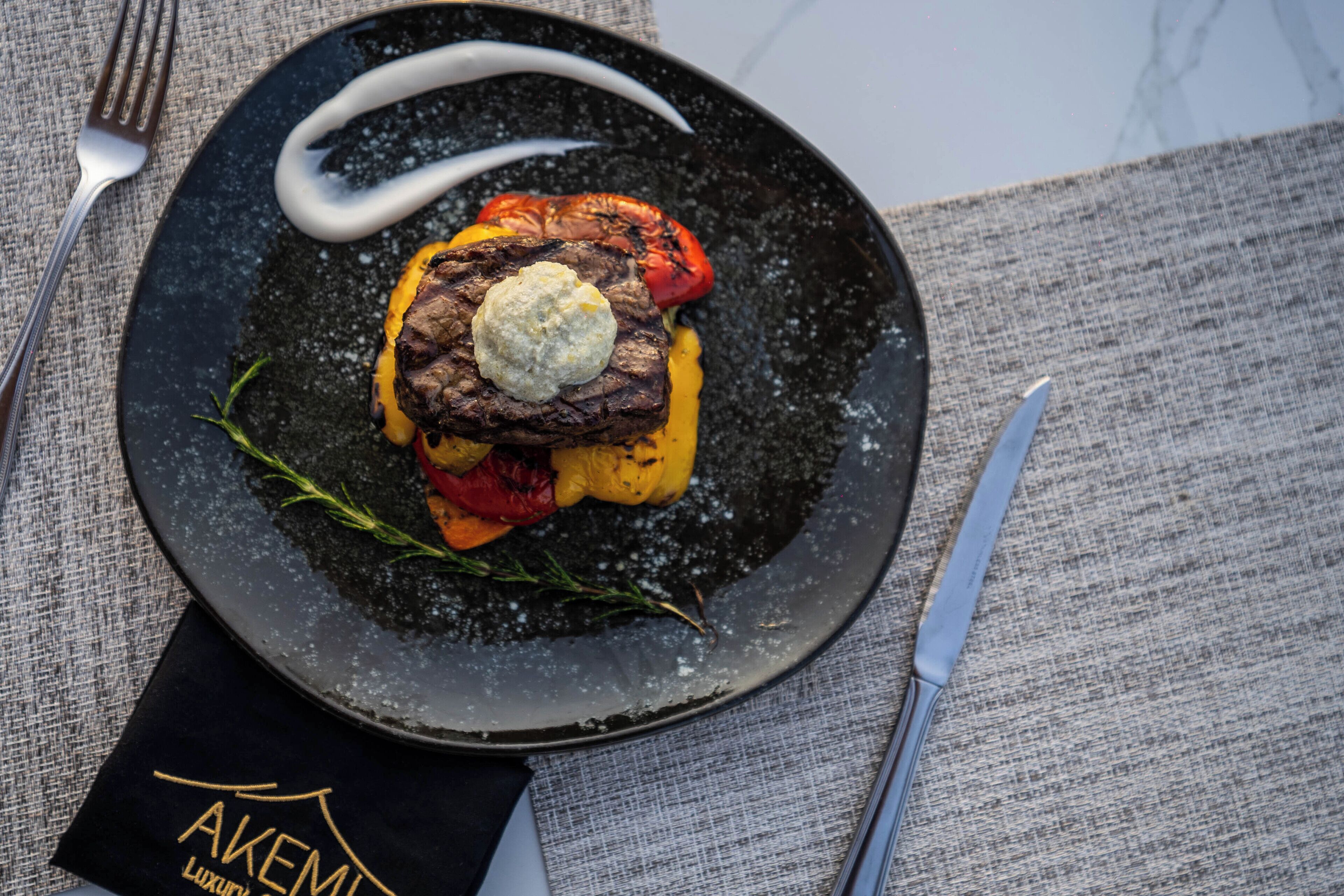 A plate of food with a steak, vegetables, and a dollop of sauce, placed on a textured tablecloth with a napkin and cutlery.
