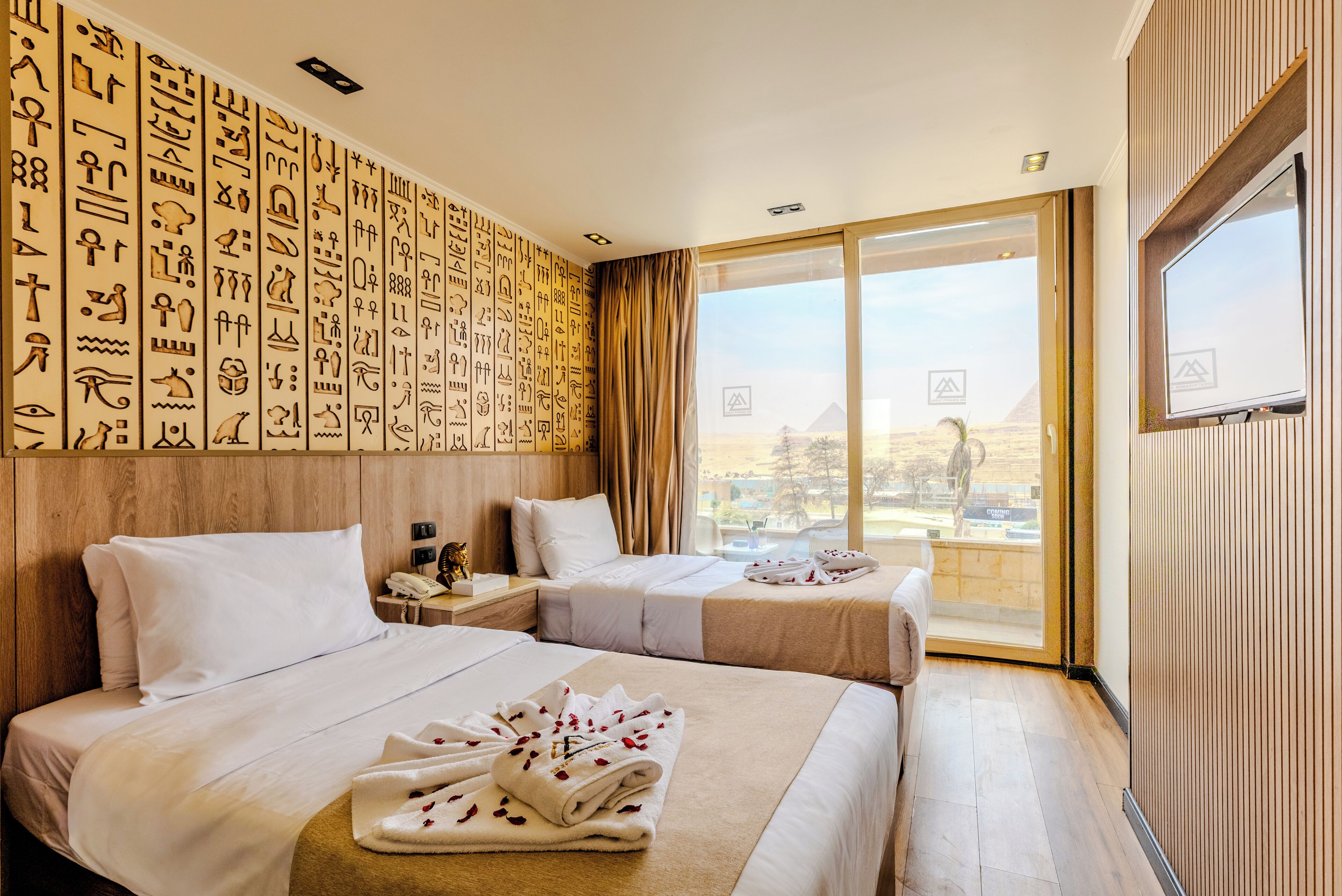 A hotel room with two beds, a large window with a view of the pyramids, a wooden wall with hieroglyphs, and a flat-screen TV.