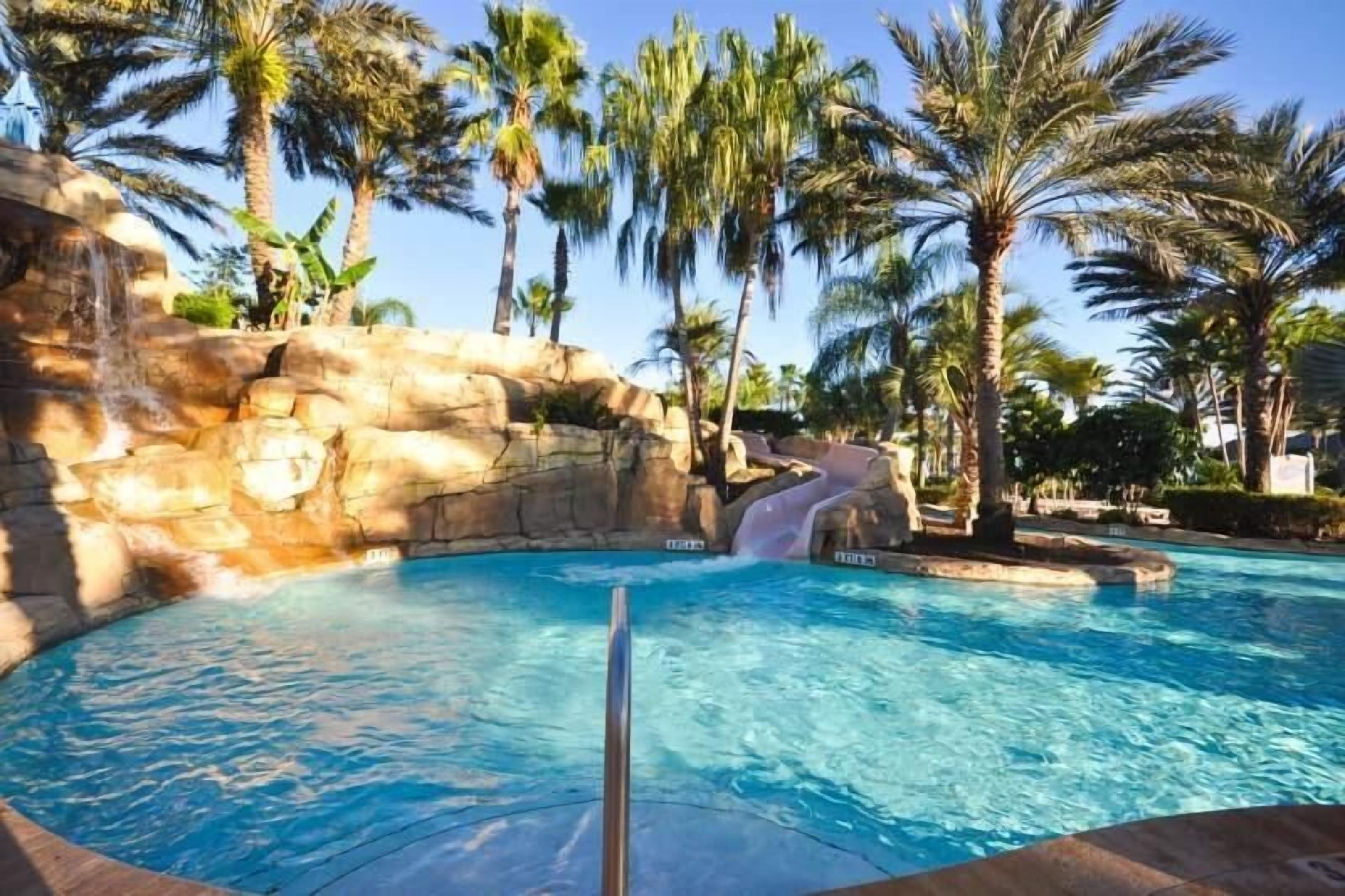 A pool with clear blue water, surrounded by palm trees and a rocky waterfall feature.