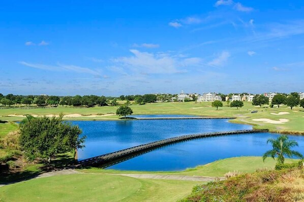 Un campo de golf con un estanque, un puente y un complejo de edificios a lo lejos.