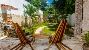 Une terrasse extérieure avec deux chevalets en bois et une petite table, entourée d’un jardin avec un sentier menant à un bâtiment blanc.