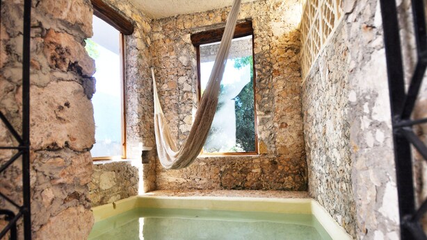 Une pièce aux murs de pierre, avec une petite piscine, deux fenêtres et un hamac.