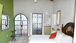 Une chambre à coucher avec un lit, une tête de lit en bois, une table de chevet ornée d’un vase de fleurs et une grande fenêtre agrémentée d’une grille en fer décorative.