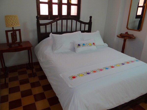 Una cama con ropa de cama blanca y almohadas florales de colores, una mesita de noche de madera con una lámpara, y un suelo a cuadros.