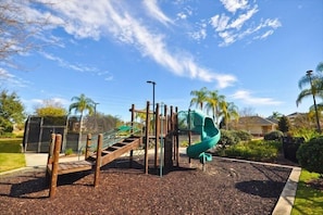 A playground with a slide and climbing structures, surrounded by a landscaped garden and residential buildings.