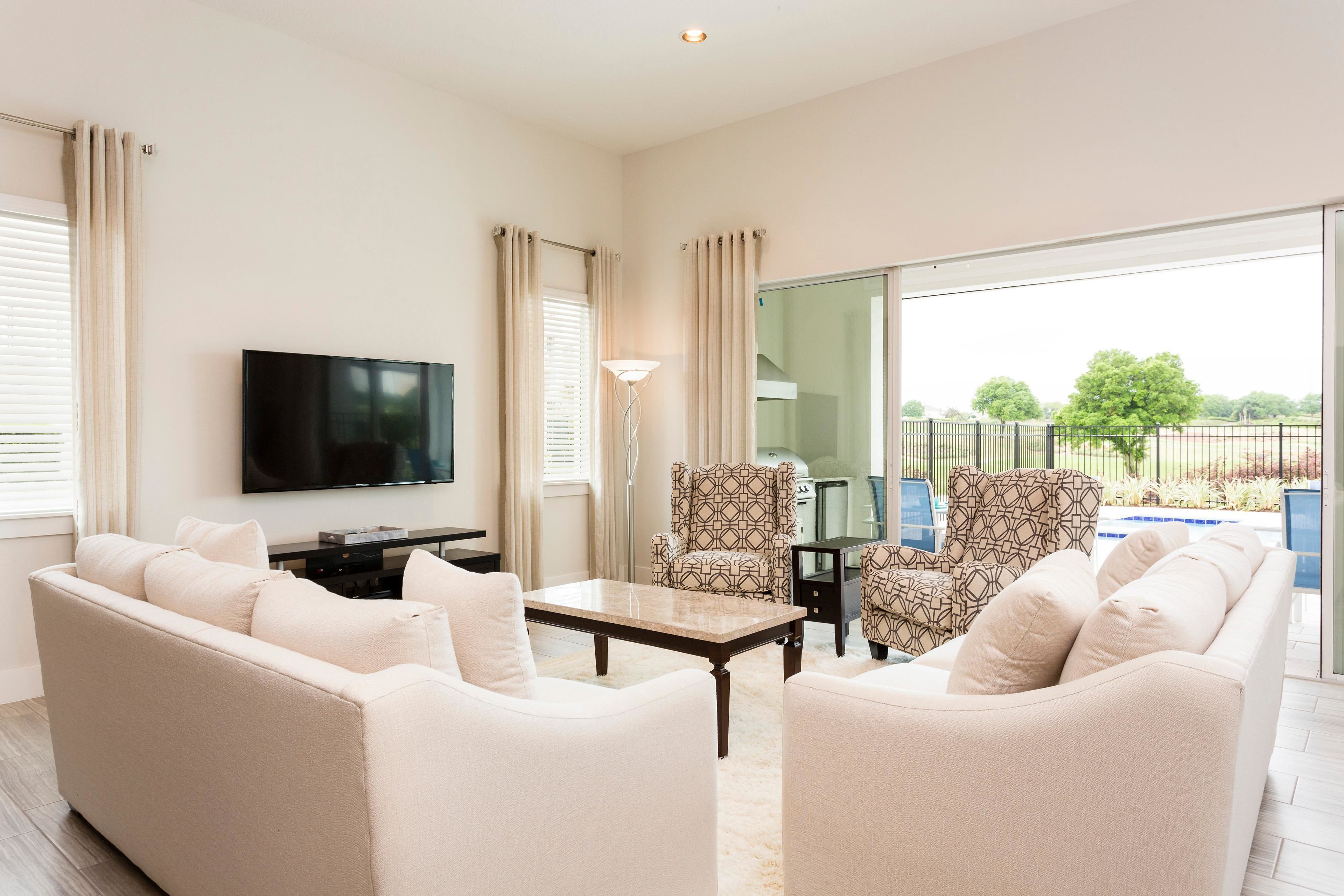 A living room with a sofa, armchairs, a coffee table, a TV, and a balcony with a pool view.