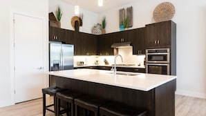 Une cuisine moderne avec des meubles de couleur bois foncé, un grand îlot central doté de tabourets, des appareils électroménagers en acier inoxydable et des plantes décoratives.