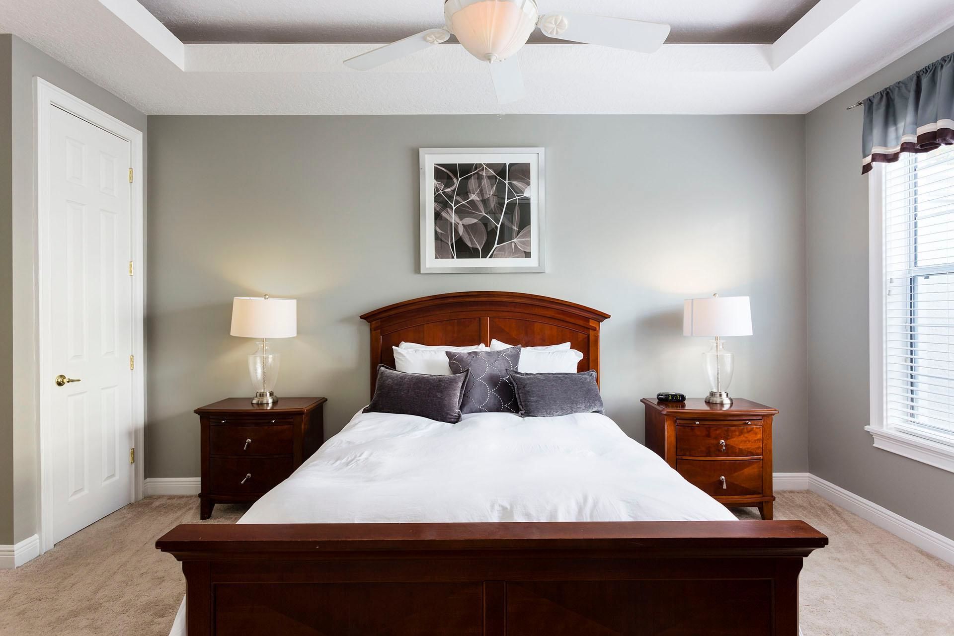 A bedroom with a large bed, two bedside tables, a ceiling fan, a framed artwork, and a window with curtains.