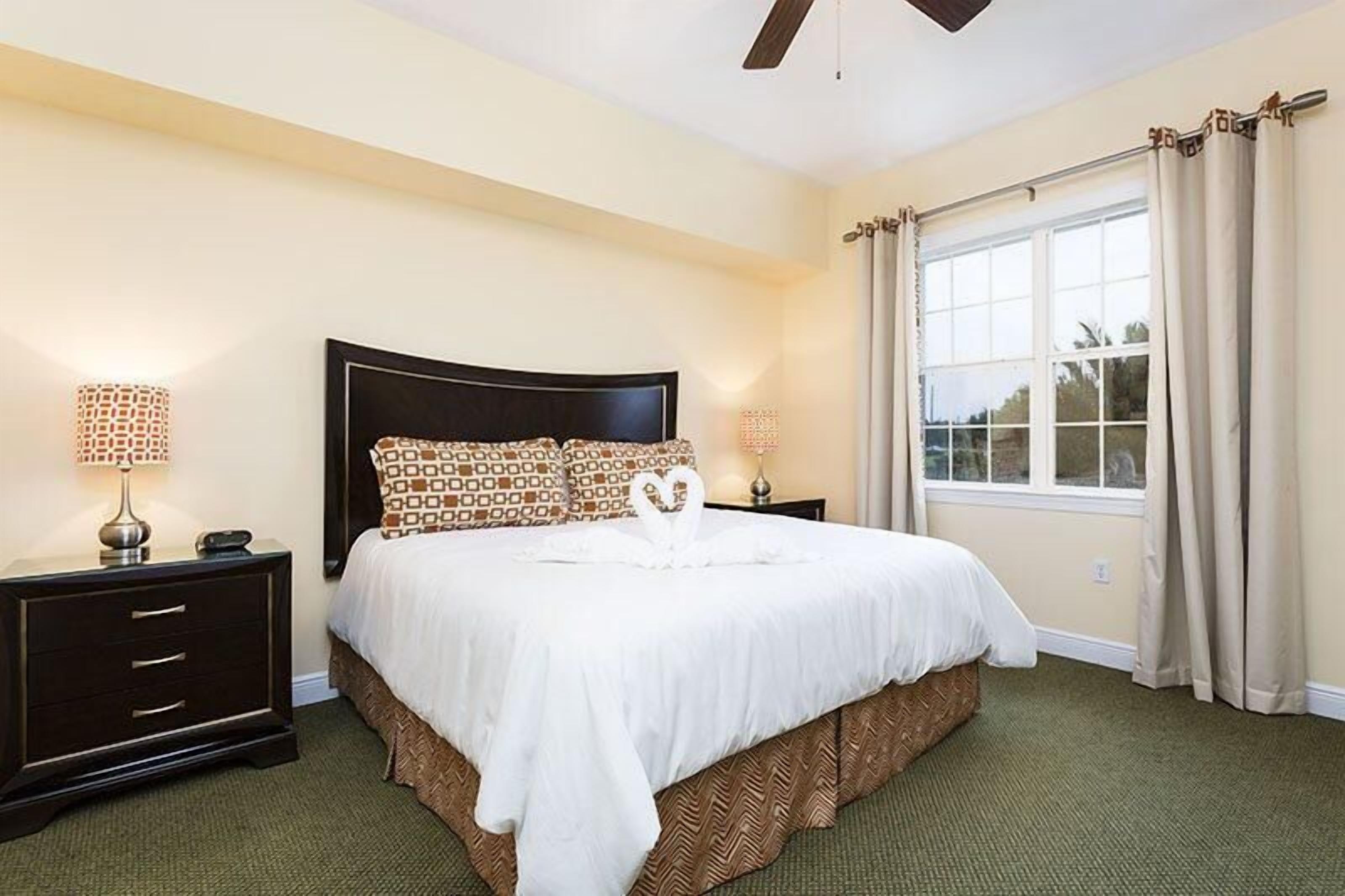 Une chambre d’hôtel avec un grand lit, deux tables de chevet, un ventilateur de plafond et une fenêtre avec des rideaux.