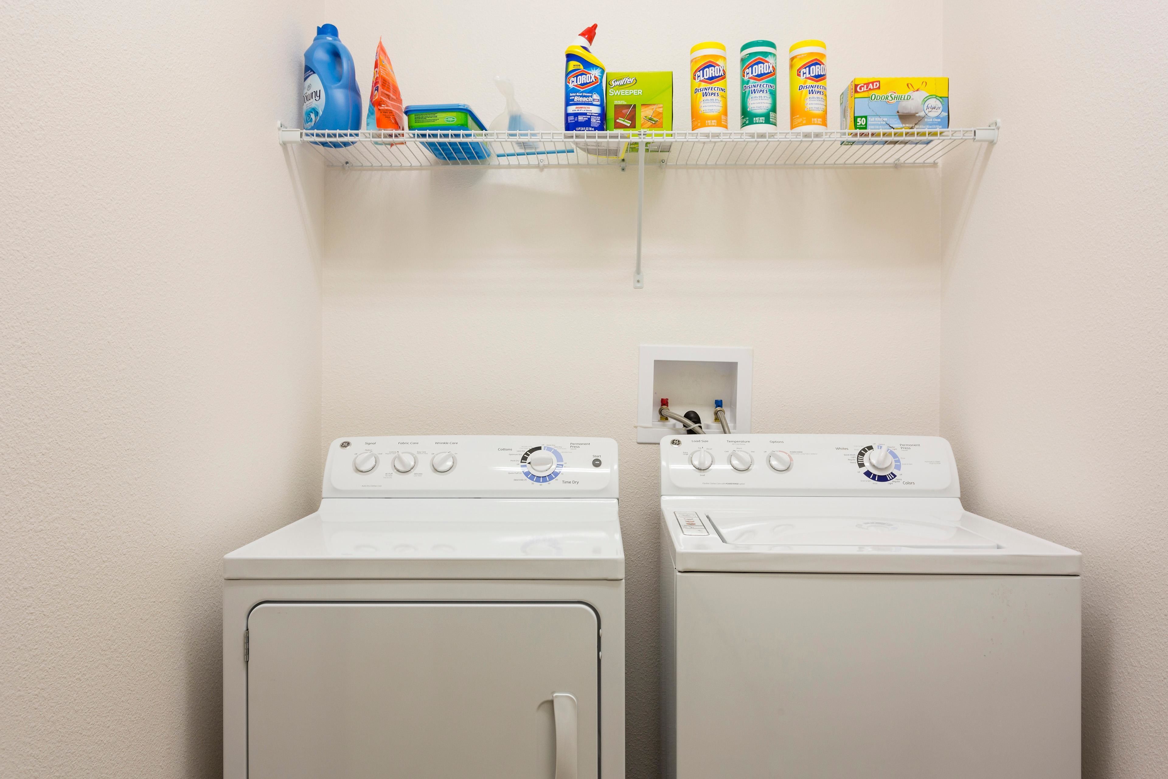 Two white front-loading washing machines with control panels, a shelf above containing cleaning products, and a small white cabinet with a red object inside.
