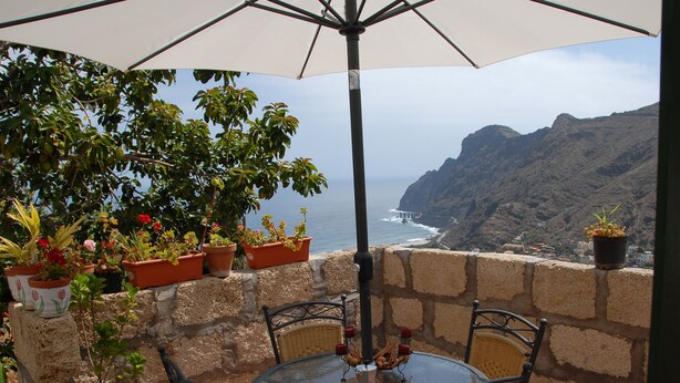 A patio with a round glass table and wicker chairs, overlooking a coastal cliff and ocean. - Casa Rural Sofia (Hermigua)