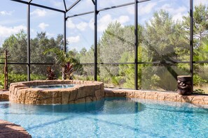 An indoor swimming pool with a natural rock feature and a waterfall, surrounded by glass walls and a view of trees.