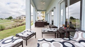 Une terrasse extérieure couverte, aménagée avec des meubles en osier, de grandes fenêtres et offrant une vue sur un terrain de golf.