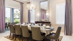 Une salle à manger avec une table ronde dressée pour six personnes, entourée de six chaises ornées de coutures décoratives. Un grand lustre est suspendu au-dessus de la table, et un miroir est fixé au mur.