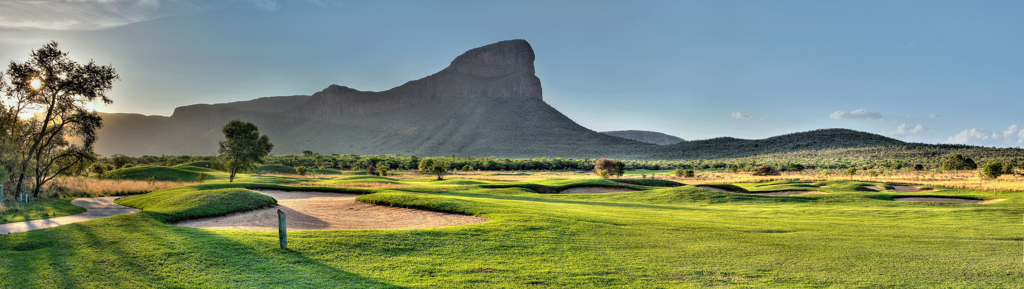 Un campo de golf con extensas zonas verdes, bunkers de arena y una montaña destacada al fondo.
