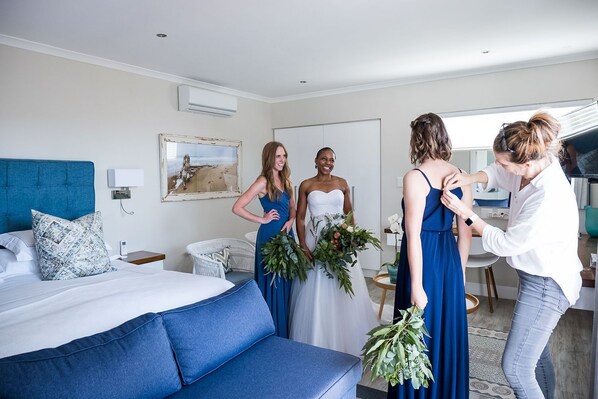 Habitación de hotel con cama, sofá azul, pared blanca con aire acondicionado, cuadro enmarcado y ventana.