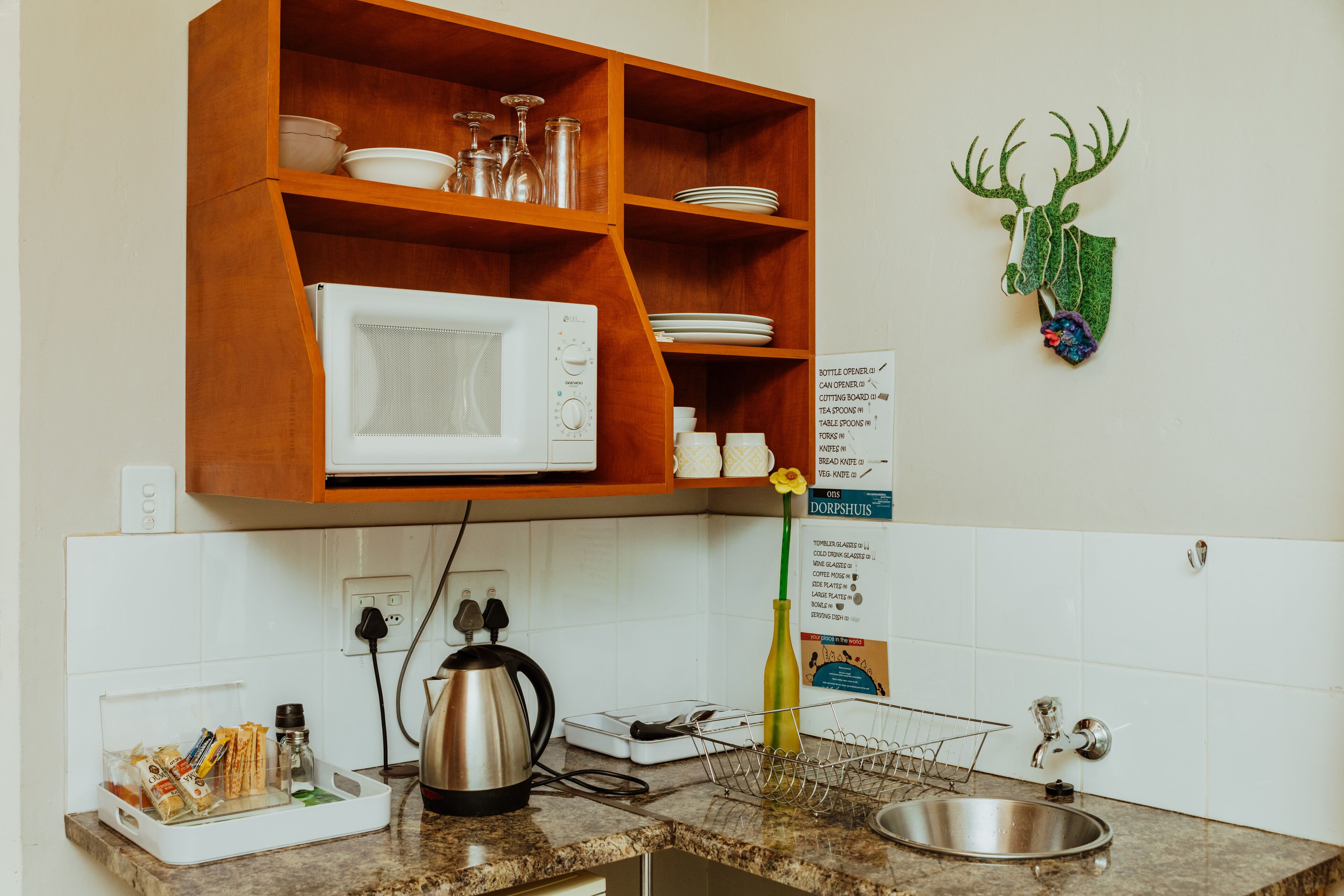 A kitchen with a microwave, shelves with dishes, a sink, and a decorative deer head.