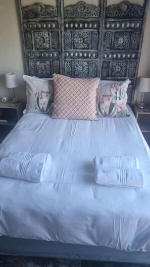 A bed with a decorative headboard, two pillows with floral patterns, and two bedside lamps. - Bluewater Reservations (Gqeberha (Port Elizabeth))