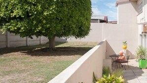 A residential building with a patio area, a dining table with chairs, a potted plant, and a large tree. - Bluewater Reservations (Gqeberha (Port Elizabeth))