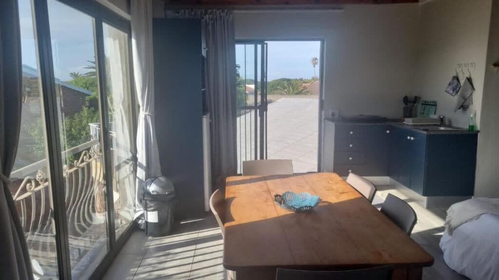 A room with a wooden table, a chair, a bed, a kitchenette, and a sliding glass door leading to a balcony.