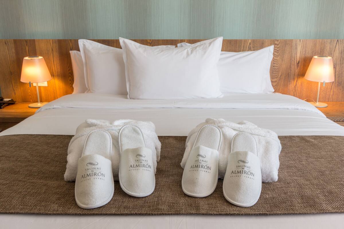 A neatly made bed with white linens and pillows, two bedside lamps, and a pair of slippers on a brown mat.