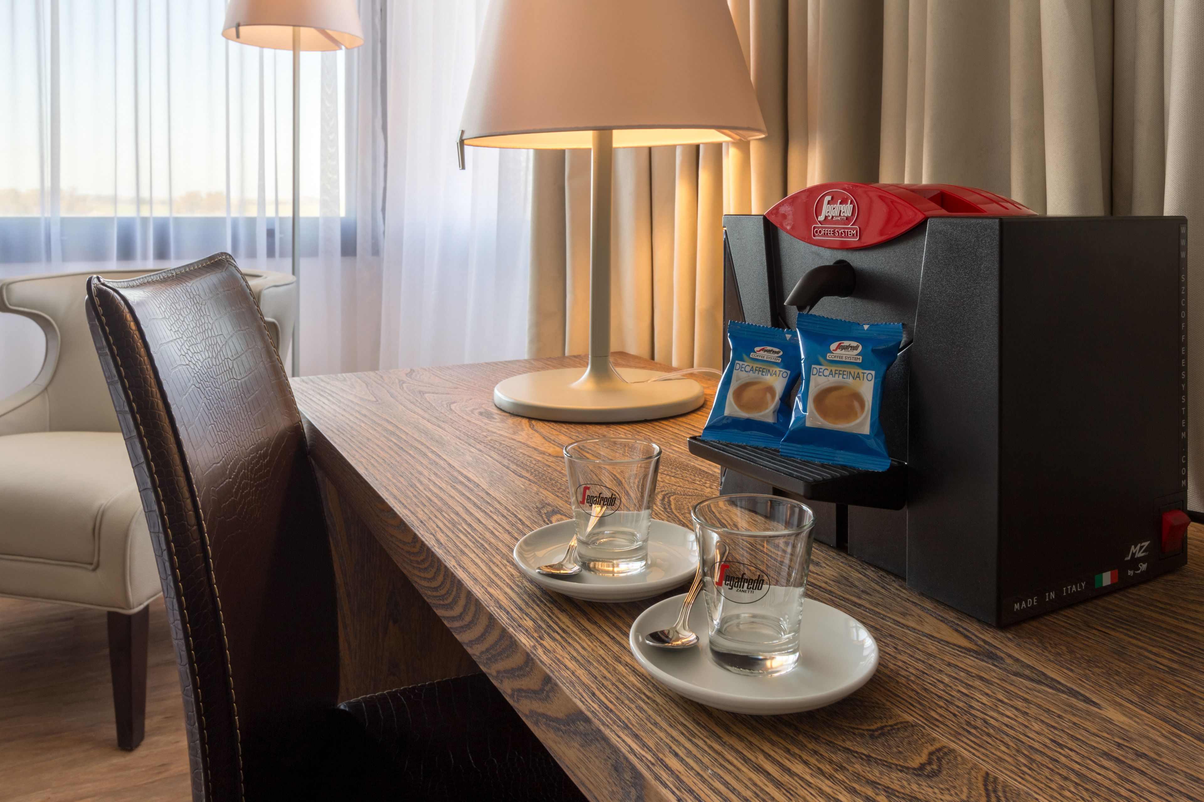 Habitación de hotel con una mesa de madera, dos vasos de agua, una lámpara y una máquina de café.