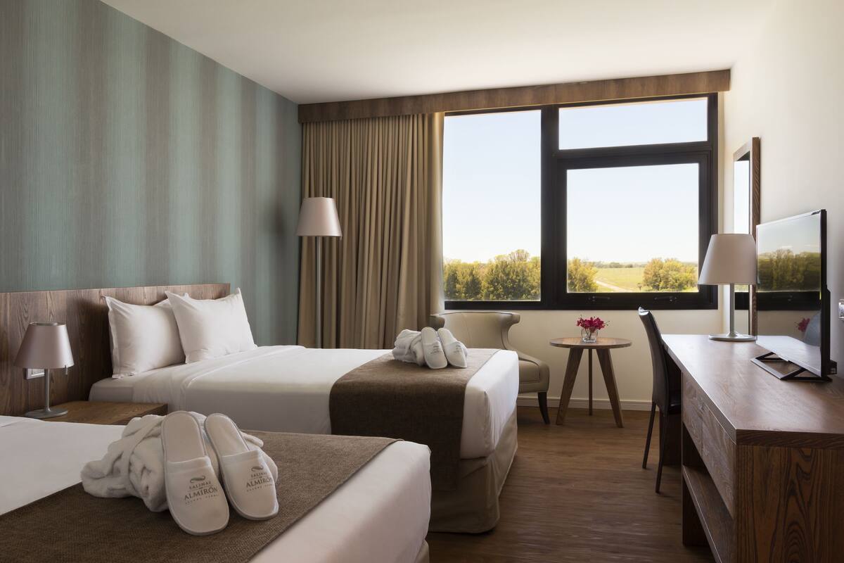 A hotel room with two beds, a desk, a chair, a small table, and a TV.